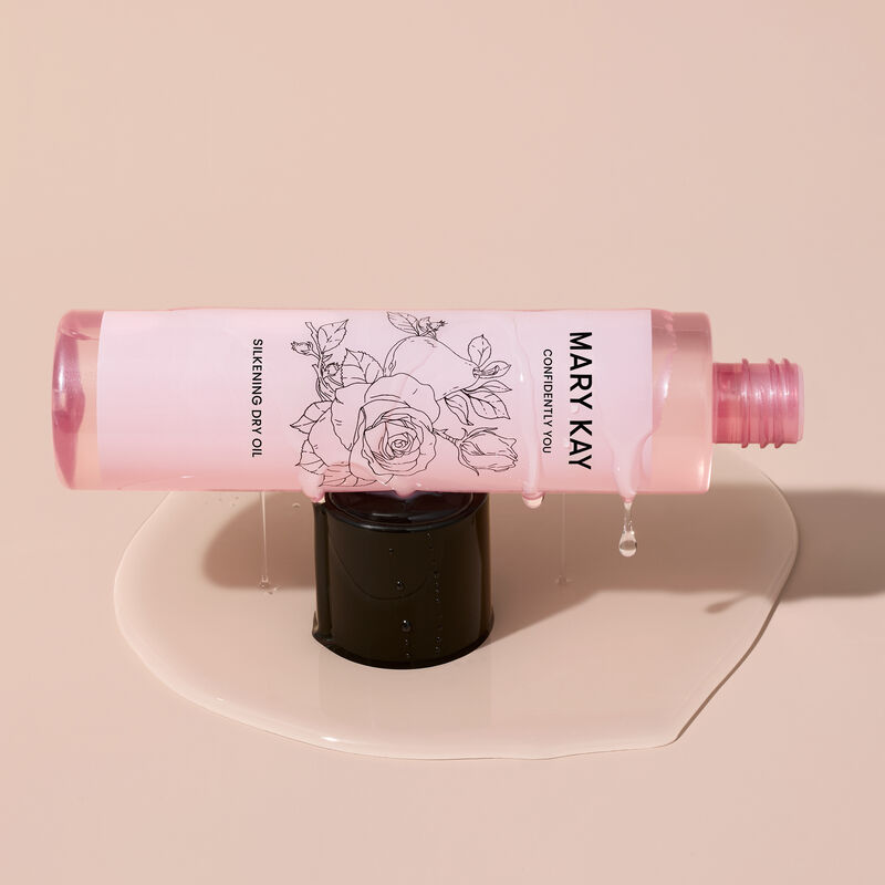 Die rosafarbene Confidently You&reg; Silkening Dry Oil Flasche liegt auf der schwarzen Verschlusskappe, die in einer kleinen Lache Trocken&ouml;l steht