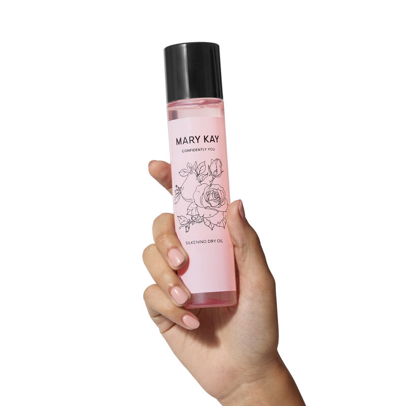 Eine Hand mit hellem Hautton h&auml;lt die rosafarbene Confidently You&reg; Silkening Dry Oil Flasche mit einer Rosen-Illustration unter dem Mary Kay Logo und 