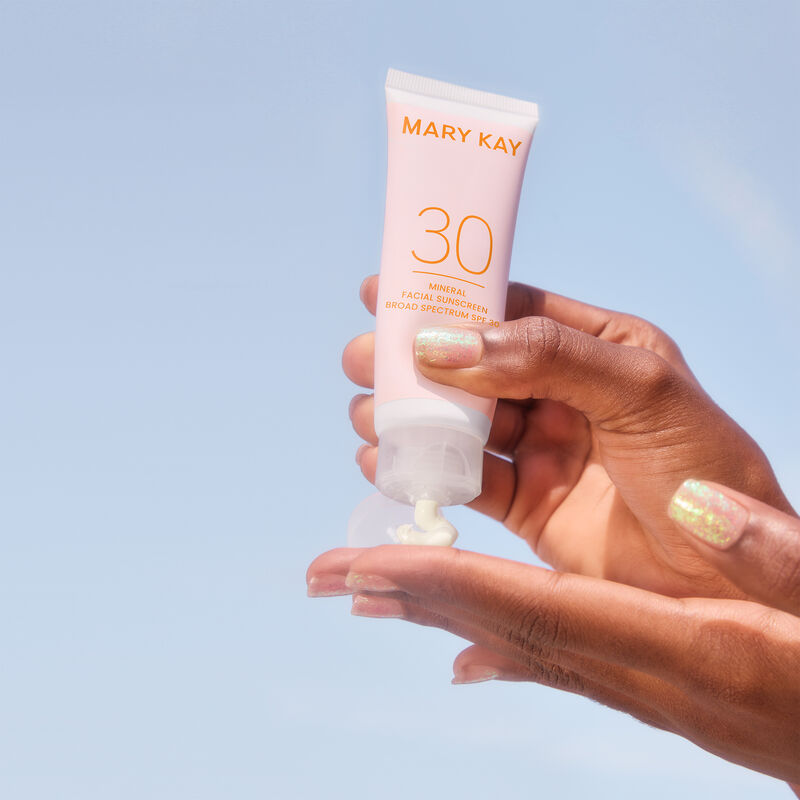 Pair of hands with deep skin tone and glitter nails squeezing product from a bottle of Mary Kay Mineral Facial Sunscreen Broad Spectrum SPF 30 image number null