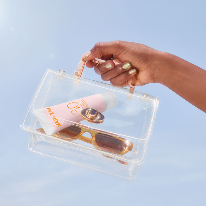 Hand with deep skin tone and glitter nails holding a small clear purse containing bottle of Mary Kay Mineral Facial Sunscreen Broad Spectrum SPF 30 an image number null