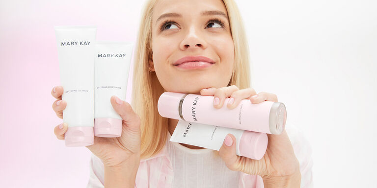 Eine l&auml;chelnde, junge Frau mit blondem Haar h&auml;lt in ihren H&auml;nden vier Produkte aus der Mary Kay Skin Care Pflegeserie f&uuml;r Mischhaut bis fettige Haut: Cleanser, Moisturizer, Peeling und Gesichtswasser
