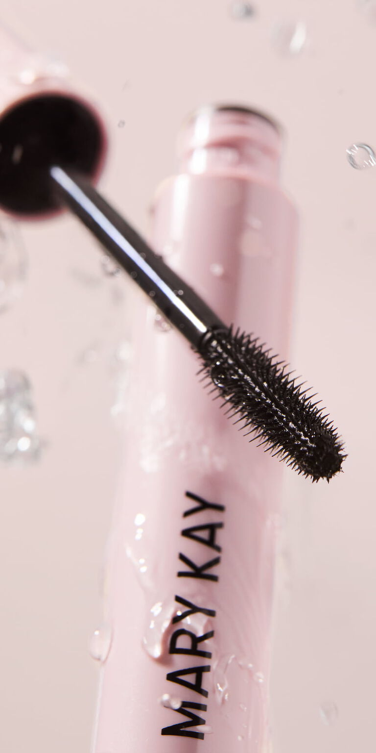 Nahaufnahme der Mary Kay Great Heights Waterproof Mascara-B&uuml;rste. Im Hintergrund sind Wassertropfen zu sehen und die ge&ouml;ffnete rosa Tube der Wimperntusche.