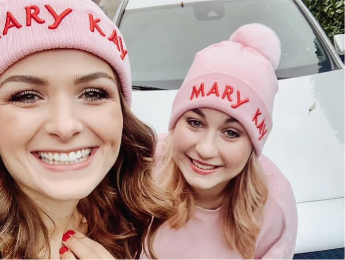 Zwei Mary Kay Beauty Consultants strahlen in die Kamera. Sie tragen rosa Mary Kay M&uuml;tzen und stehen vor einem Mercedes, den sie sich durch ihre T&auml;tigkeit als Beauty Consultant erarbeitet haben.