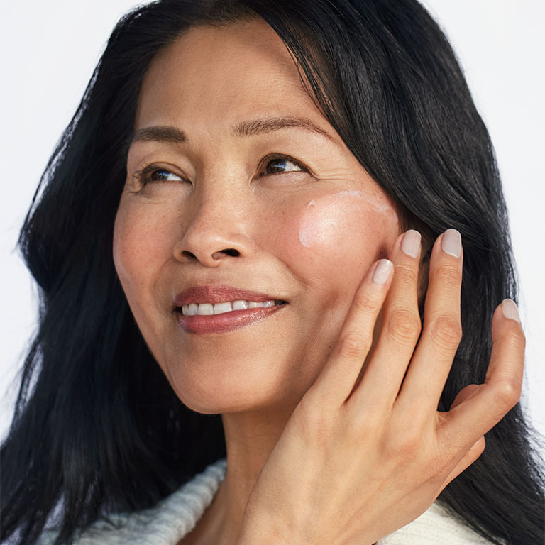 Eine l&auml;chelnde Frau in einem wei&szlig;en Oberteil cremt sich das Gesicht mit Sunscreen SPF 50+ von Mary Kay ein