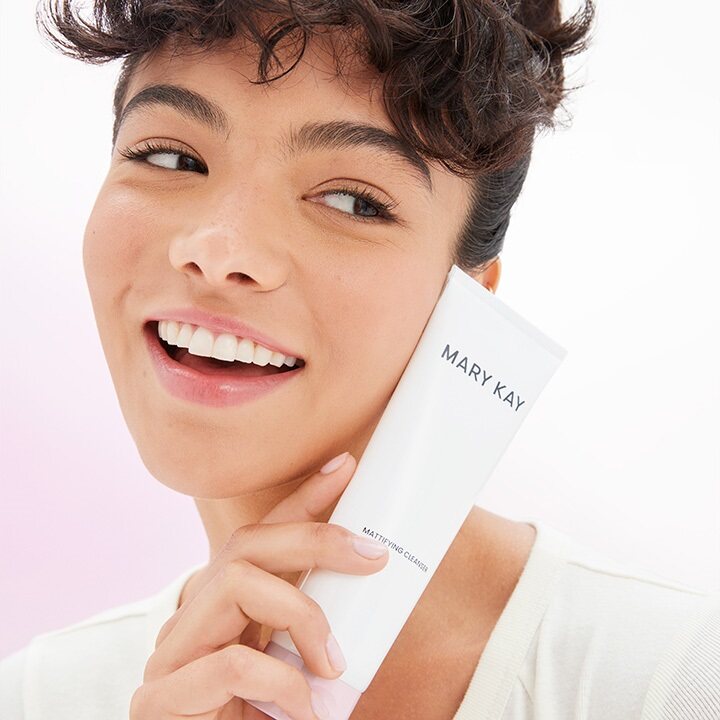 Eine junge Frau mit gepflegter Haut l&auml;chelt und h&auml;lt mit einer Hand die wei&szlig;e Tube des Mary Kay Mattifying Cleanser auf der H&ouml;he ihres Gesichts