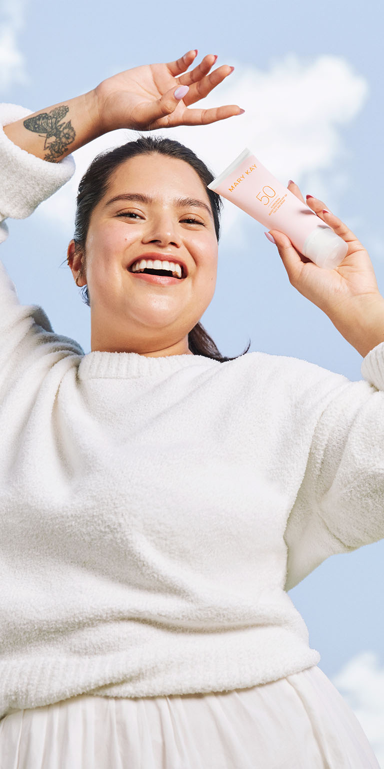 Eine l&auml;chelnde, in wei&szlig; gekleidete Frau lacht, streckt die Arm in die Luft und h&auml;lt in einer Hand die rosa Tube der Mary Kay Sonnencreme mit Lichtschutzfaktor 50+. Hinter ihr ist ein strahlend blauer Himmel mit kleinen wei&szlig;en Wolken zu sehen.