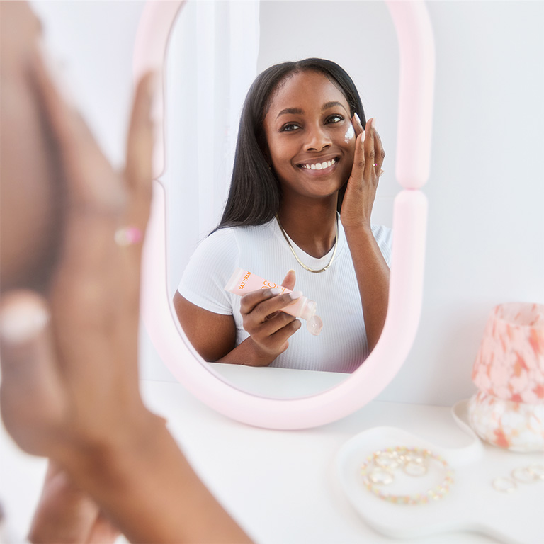 Auf einem wei&szlig;en Tisch steht ein rosa Spiegel, in dem man sieht, wie eine Frau sich mit einer Hand das Gesicht mit dem Mary Kay Mineral Facial Sunscreen LSF 30 Sonnenschutz eincremt. In der anderen Hand h&auml;lt sie die Sonnencreme Tube.